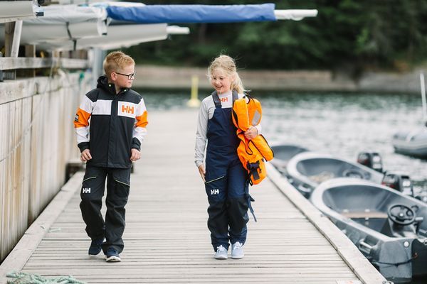 Två barn går längs en brygga vid vattnet, klädda i sjökläder med Helly Hansen-logotyp. Pojken till vänster bär en mörk jacka med orange och vita detaljer, medan flickan till höger håller en orange flytväst och bär en blå overall. Båtar ligger förtöjda vid bryggan, och i bakgrunden syns en skogskant