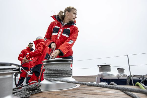 En grupp människor klädda i röda seglarjackor står ombord på en båt och arbetar med riggen. Kvinnan i förgrunden hanterar en vinsch med ett rep, medan två andra besättningsmedlemmar arbetar i bakgrunden. Himlen är grå och vädret verkar mulet, vilket antyder att de befinner sig till sjöss under en seglingstur.